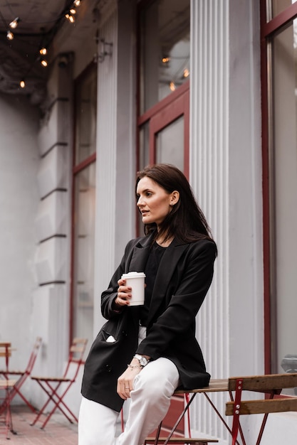 Zelfverzekerde jonge zakenvrouw met kopje koffie poseert buiten café op terras Vrolijk meisje draagt casual zakelijke stijl poseert in de buurt van café