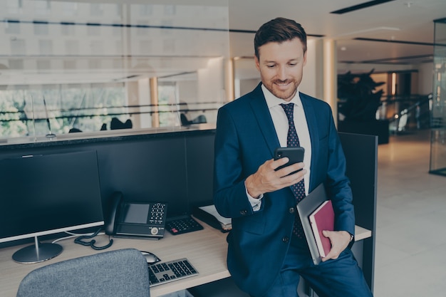 Zelfverzekerde lachende ondernemer die smartphone gebruikt en glimlacht terwijl hij op kantoor staat