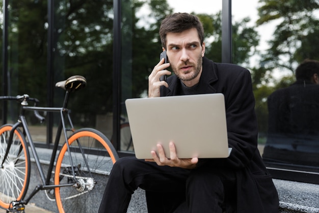 Zelfverzekerde man gekleed in jas zittend aan de straat, met behulp van laptopcomputer, praten op mobiele telefoon