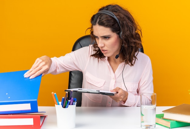 Zelfverzekerde mooie blanke vrouwelijke callcenter-operator op koptelefoon zittend aan een bureau met kantoorhulpmiddelen met klembord en kijken naar bestandsmap geïsoleerd op oranje muur