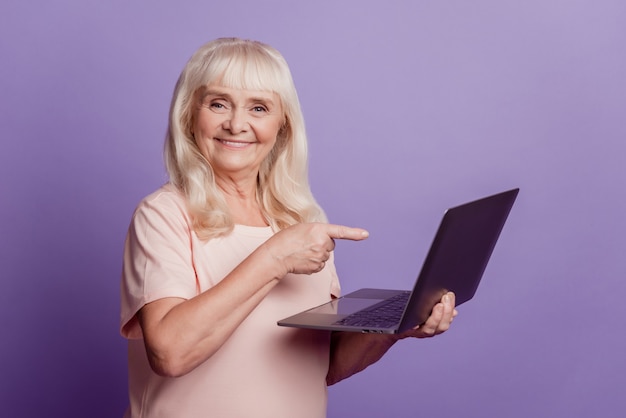 Zelfverzekerde oudere vrouw houdt punt vinger laptop geïsoleerd op violette achtergrond