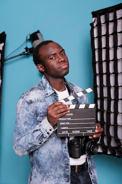 Zelfverzekerde producer in studio met filmklapper terwijl hij naast professionele productieapparatuur staat. Fotograaf met klepel op blauwe achtergrond naast moderne schijnwerpers en softboxen.