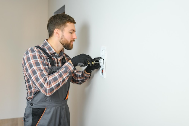 Foto zelfverzekerde professionele elektricien in uniform die een schroevendraaier gebruikt tijdens het vervangen van een stopcontact in een appartement na renovatiewerkzaamheden