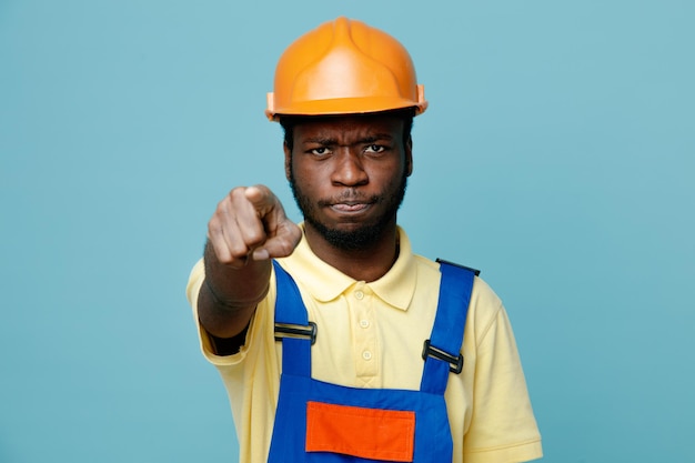Zelfverzekerde punten op camera jonge afro-amerikaanse bouwer in uniform geïsoleerd op blauwe achtergrond
