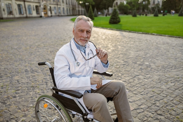 Foto zelfverzekerde senior gehandicapte mannelijke arts in rolstoel met laboratoriumjas en bril kijkend naar