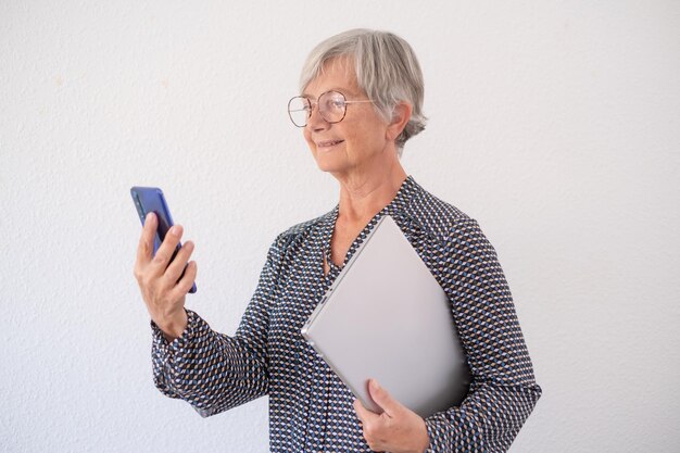 Zelfverzekerde senior vrouw in shirt geïsoleerd op een witte achtergrond met laptop onder haar arm tijdens het gebruik van mobiele telefoon