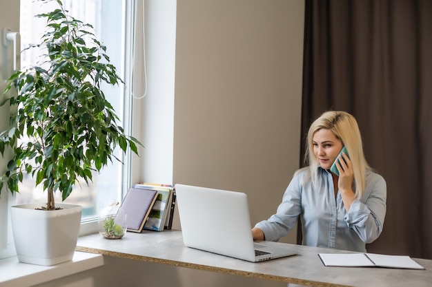 Zelfverzekerde serieuze ervaren gekwalificeerde mooie slimme vrouw met blond haar die shirt draagt, typt brieven aan haar klanten en zakenpartners, zittend aan de tafel in kantoor