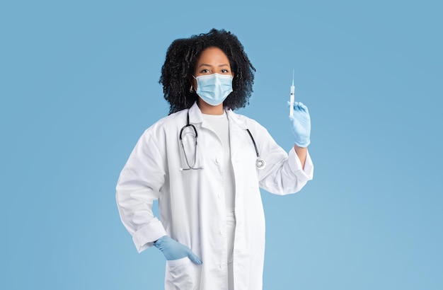 Zelfverzekerde serieuze jonge zwarte dame arts in witte jas beschermend masker en handschoenen toont spuit
