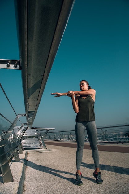 Zelfverzekerde sportvrouw op de brug die arm opzij legt terwijl ze haar schouderspieren opwarmt