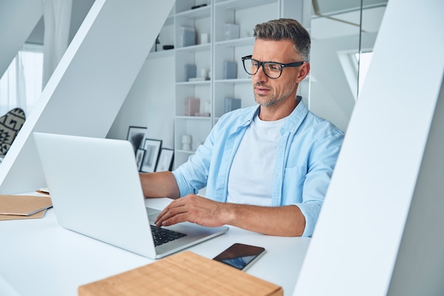 Zelfverzekerde volwassen man die op laptop werkt terwijl hij aan het bureau zit