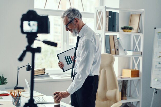 Zelfverzekerde volwassen man in elegant overhemd en stropdas die iets vertelt tijdens het maken van video op sociale media