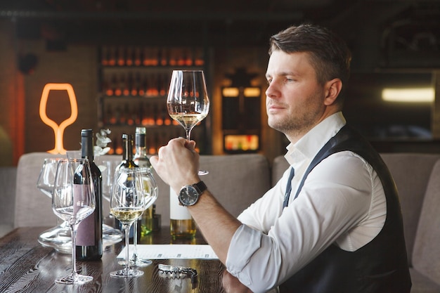 Zelfverzekerde volwassen man sommelier kijkt naar glanzend wijnglas dure witte wijn zittend aan tafel in luxe restaurant close-up wijndegustatie