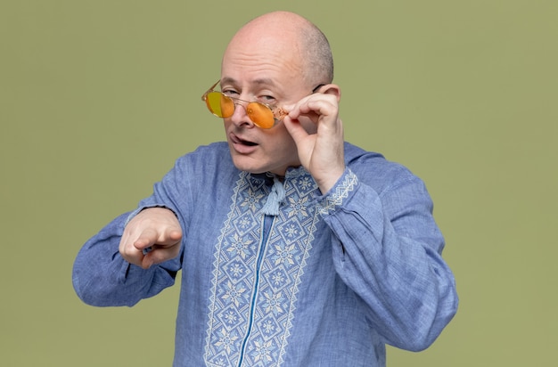 Zelfverzekerde volwassen Slavische man in blauw shirt met een zonnebril die naar de camera kijkt en wijst