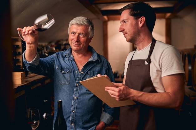 Zelfverzekerde volwassen sommelier die een glas rode wijn kantelt en ernaar kijkt met zijn assistent