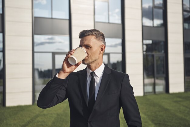 Zelfverzekerde volwassen zakenman die koffie drinkt terwijl hij in de buurt van een kantoorgebouw staat