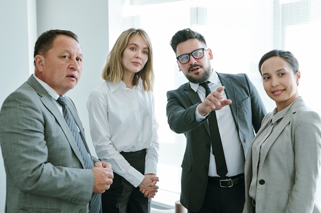 Zelfverzekerde volwassen zakenman die naar voren wijst terwijl hij tussen interculturele collega's staat tijdens een discussie tijdens een vergadering op kantoor