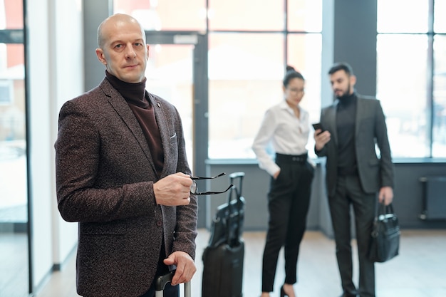 Zelfverzekerde volwassen zakenman met bril en koffer staan met twee jonge collega's