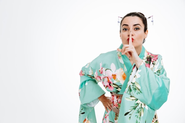 Zelfverzekerde vrouw in traditionele Japanse kimono die stiltegebaar met vinger op lippen op wit maakt