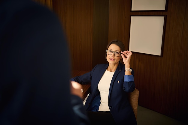 Zelfverzekerde vrouw van middelbare leeftijd die streeft naar zakelijk succes zit ontspannen in een fauteuil in een hotelkamer en praat terwijl ze naar haar zakenpartner kijkt. Kaukasische vrouwen die zakelijk succes behalen