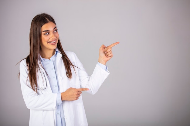 Foto zelfverzekerde vrouwelijke arts in wit uniform wegkijken en wijzen terwijl staande tegen een grijze achtergrond. kijk daar! dit is wat uw arts aanbeveelt.