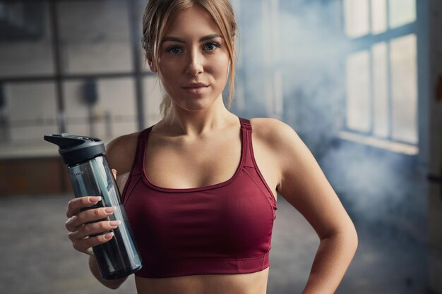 Zelfverzekerde vrouwelijke atleet in sportkleding en met een fles verfrissend water kijkend naar de camera in de sportschool met rook