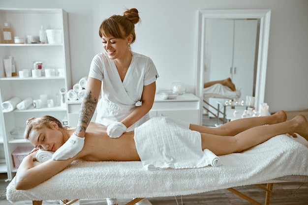 Zelfverzekerde vrouwelijke specialist doet ontspannende lichaamsmassage voor mooie slanke blanke dame in minimalistische spa-salon