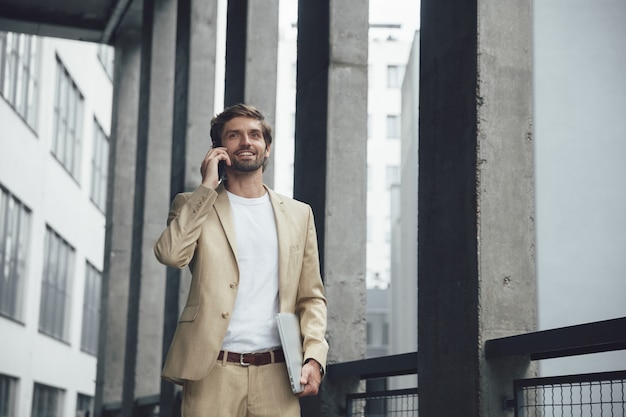 Zelfverzekerde zakenman in formele outfit praten op mobiel terwijl buitenshuis staan met laptop in de hand
