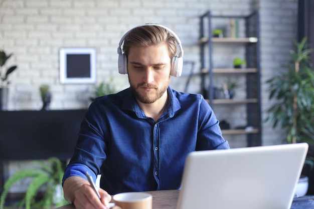 Zelfverzekerde zakenman in oortelefoons schrijft notities of financieel rapport terwijl hij thuis aan een bureau zit met een laptop.