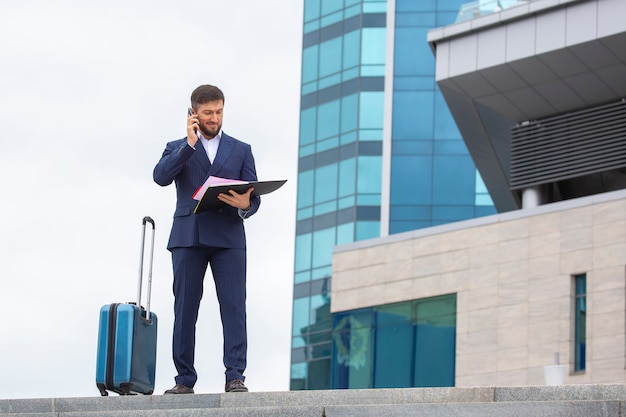 Zelfverzekerde zakenman staat met een reiskoffer op de trap tegen de achtergrond van een kantoorgebouw