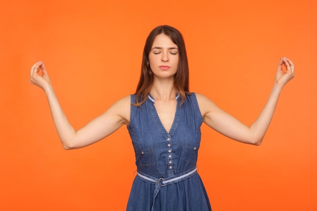 Zen praktijk adem techniek brunette vrouw in denim jurk doen yoga oefening hand in hand in mudra gebaar en mediteren gevoel rustige ontspannen studio opname geïsoleerd op oranje achtergrond