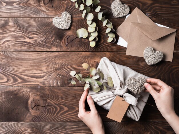 Zero waste Valentine's Day eco-friendly gift wrapping in Furoshiki style with dry eucalypt.Hands with gift box in cloth wrapping with empty craft label on wooden background.Copy space.Topview, flatlay