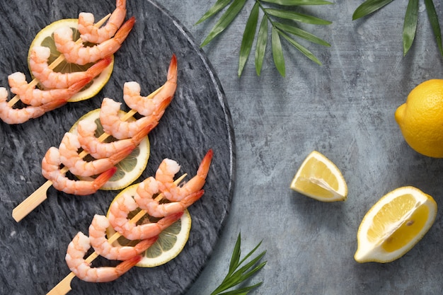 Zesty lemon and garlic prawns on wooden sticks, flat lay on dark stone