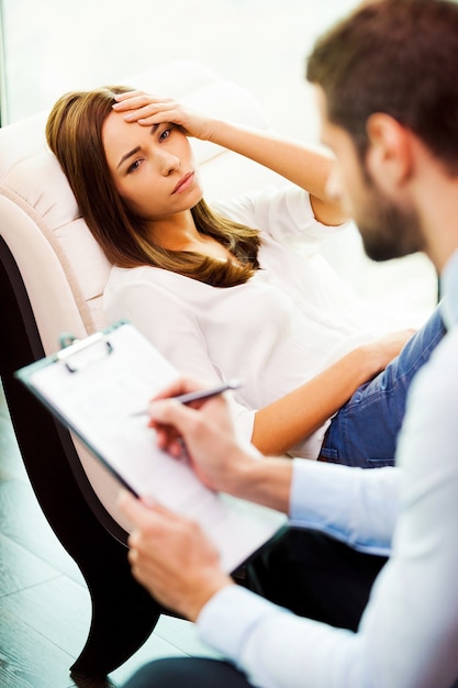 Zich hopeloos en depressief voelen. Depressieve jonge vrouw zit aan de stoel en houdt de hand op het hoofd terwijl een jonge man dicht bij haar zit en iets in zijn klembord schrijft