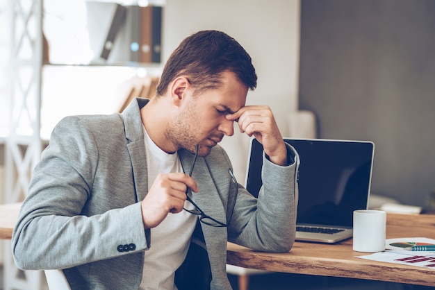 Zich moe voelen. Gefrustreerde jonge knappe man die er uitgeput uitziet terwijl hij op zijn werkplek zit en zijn bril in de hand draagt