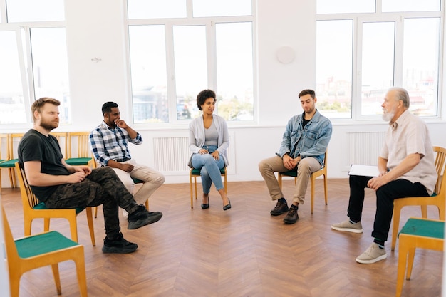 Zicht van een zelfverzekerde volwassen mannelijke coachpsycholoog die in een cirkel zit tijdens groepstherapie met mensen van verschillende leeftijden.