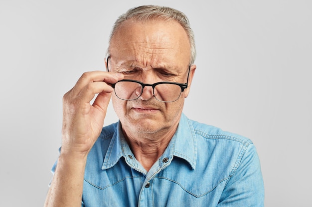 Zichtproblemen. Een oudere man zet zijn bril af. Niet tevreden. Senior burger op een witte achtergrond in een blauw shirt. Optiek