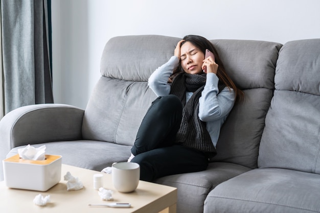 Ziek Aziatisch meisje dat thuis op de bank in de woonkamer zit en met de arts of haar familie op de mobiele telefoon praat voor counseling, vrije ruimte. Verkoudheid, griep, coronavirus pandemie of thuisisolatie concept.