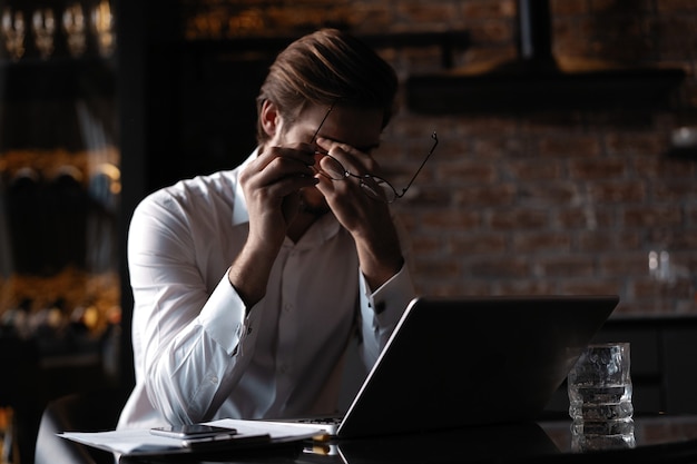 Ziek en moe voelen. Gefrustreerde jongeman die zijn neus masseert en zijn ogen gesloten houdt terwijl hij op een laptop in café zit.