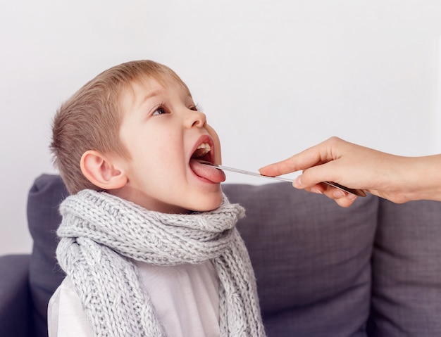 Ziek kind bevriest verpakt in een sjaal. De moeder van een kleine jongen gebruikt een lepel om de keel te onderzoeken