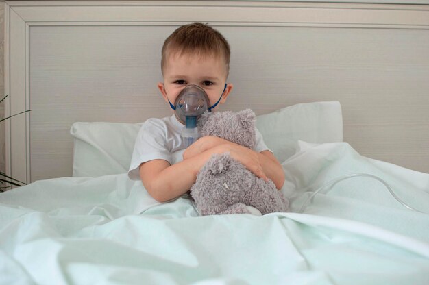 Ziek kind is ziek thuis, liggend in bed met teddybeer. voert de inhalatieprocedure uit met een vernevelaar van de luchtwegen. behandeling van een kind voor hoest. compressie inhalator. selectieve aandacht.