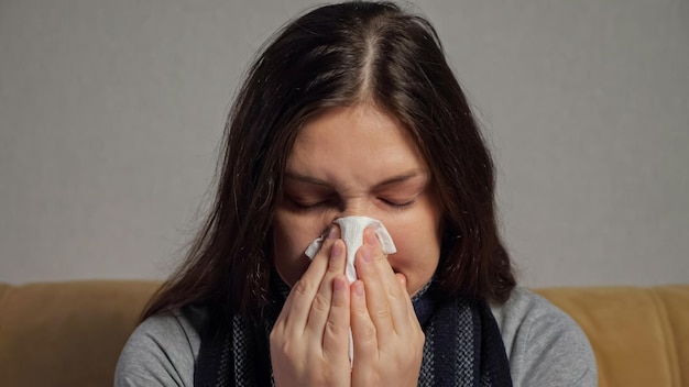 Zieke depressieve brunette vrouw gewikkeld in sjaal met loopneus niest in servet zittend onder op de bank die lijdt aan griep, close-up