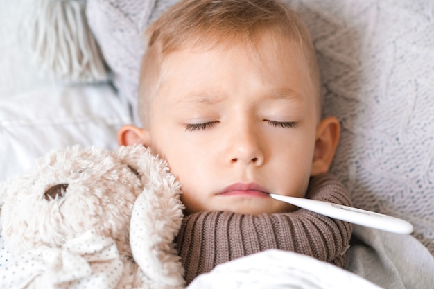 Zieke en verdrietige jongen ligt in bed met een thermometer in zijn mond en een zacht stuk speelgoed in zijn armen