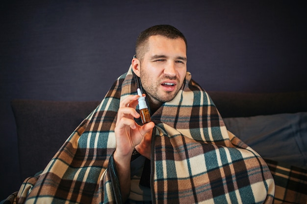 Zieke en zieke jonge man houdt neusspray in handen. Hij krimpt maar kijkt op camera. Guy gaat niezen. Hij ziet er slecht uit. Man zit in de slaapkamer.