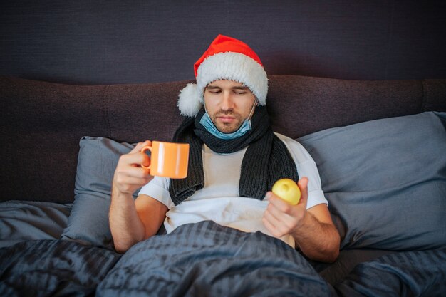 Zieke jonge man in rode kerstmuts zit op het bed