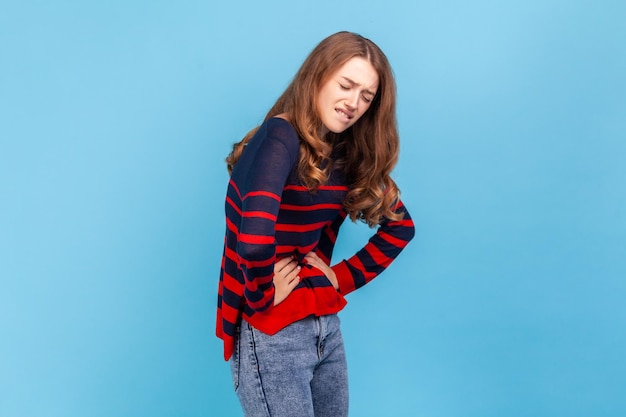 Zieke jonge volwassen vrouw die een gestreepte trui in casual stijl draagt, haar buik aanraakt, lijdt aan acute pijn buikkrampen, risico op een miskraam. Indoor studio opname geïsoleerd op blauwe achtergrond.