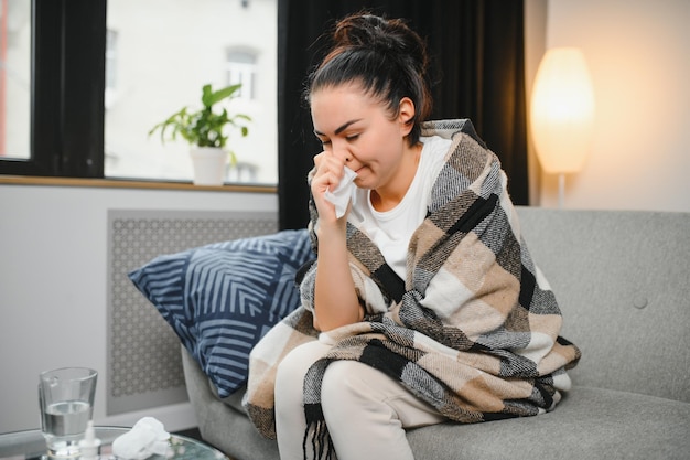 Zieke jonge vrouw zit op de bank bedekt met deken die bevriest en blaast haar loopneus niest in weefsel Vrouw kreeg koorts griep- en griepsymptomen hoesten thuis Ziek allergisch voor luchtwegaandoeningen