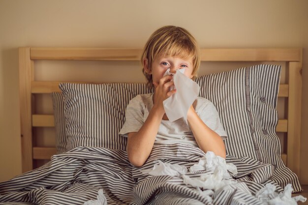 Zieke jongen hoest en veegt zijn neus af met doekjes Ziek kind met koorts en ziekte in bed