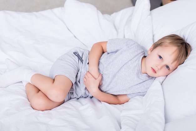 Zieke jongen liggend op bed en pijnlijke maag in slaapkamer aan te raken, camera kijken