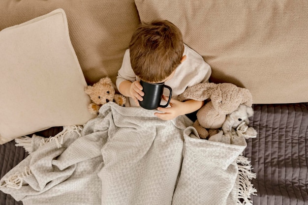 Zieke kleine jongen die thuis hete thee drinkt op het bed. Onwel, ziek kind gewikkeld in een deken, met mok in zijn kamer. Griepseizoen. Interieur en kleding in natuurlijke aardekleuren. Gezellige omgeving.