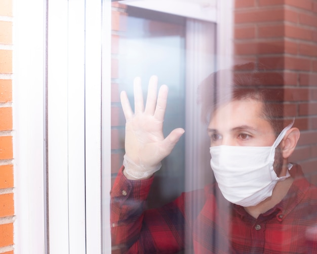 Foto zieke man met kroonvirus kijkt uit het raam en draagt een beschermend masker